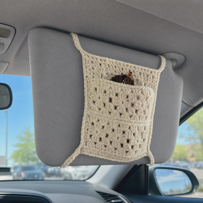 May include: A cream-colored crochet car visor organizer featuring a square granny square pattern with a central pocket. The organizer is secured to a gray vehicle sun visor with crocheted straps and contains a pair of brown sunglasses.