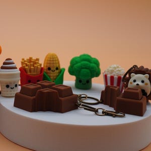 Könnte beinhalten: Eine Sammlung farbenfroher, miniaturisierter Figuren mit Essen und Tieren. Enthält einen Maiskolben, einen Drachen, eine Tomate, ein Kaninchen auf einer Karotte, einen Schokoriegel und einen Igel. Die Figuren werden auf weißen Podesten vor orangefarbenem Hintergrund präsentiert.