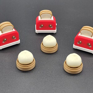 Könnte beinhalten: Eine Sammlung von Miniatur-Essensspielzeugen, darunter rote Toaster mit Brot und weiße Brötchen in Bambusdämpfern. Die Toaster haben ein lächelndes Gesichtsdesign. Die Spielzeuge sind auf einer dunkelgrauen Oberfläche angeordnet.