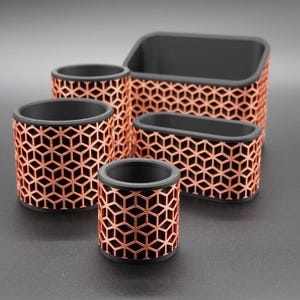 Könnte beinhalten: Ein Set aus fünf Schreibtisch-Organizern in verschiedenen Formen und Größen, mit mattschwarzem Innenraum und einer kupferfarbenen Außenseite mit einem geometrischen Gittermuster aus sich wiederholenden Würfeln. Das Set enthält drei zylindrische Behälter.