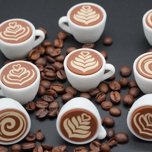 Könnte beinhalten: Acht Miniatur-Kaffeetassen aus weißer Keramik, angeordnet zwischen verstreuten gerösteten Kaffeebohnen. Jede Tasse hat eine flache braune Oberfläche mit detaillierten Latte-Art-Mustern, darunter herzförmige Designs und kreisförmige Wirbel.
