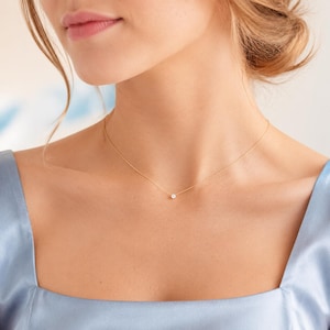 May include: A delicate gold chain necklace featuring a single, small, round, clear gemstone pendant centered on the collarbone. The necklace is worn against a light blue satin garment with a square neckline, highlighting the minimalist design.