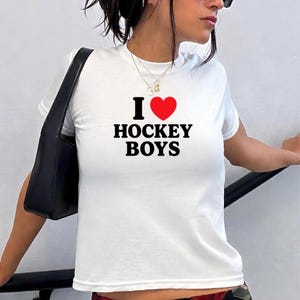 Puede incluir: Una camiseta blanca de manga corta con el texto negro "I" seguido de un símbolo de corazón rojo y el texto negro "HOCKEY BOYS" impreso en el centro del pecho. La camiseta tiene un cuello redondo y un corte estándar, junto a un bolso negro.