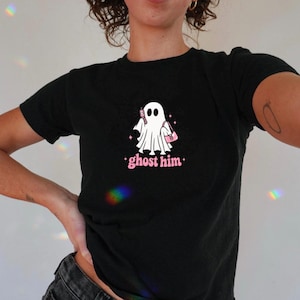 May include: A black short-sleeved t-shirt featuring a white ghost graphic holding a pink handbag and a cell phone. Below the ghost, the text reads "ghost him" in pink letters, surrounded by small pink star accents on the front of the garment.