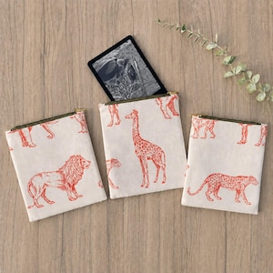 May include: Three square fabric pouches featuring a repeating orange animal print on a cream background. The designs include a lion, a giraffe, and a leopard. Each pouch has a dark green zip closure at the top. A black tablet rests on the centre pouch.