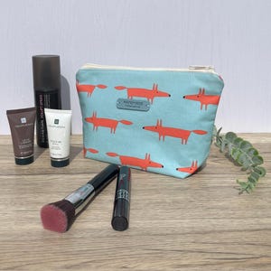 May include: A light blue zipped pouch featuring a repeating orange fox pattern and a small grey tag labelled HANDMADE. Beside it are two small tubes of TempleSpa skincare, a black spray bottle, a makeup brush with pink bristles, and a black mascara.