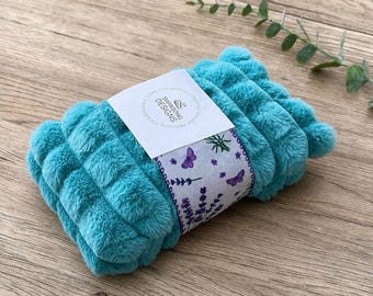 May include: A folded teal plush blanket with a grid-like textured surface. A white paper band wraps around the center, featuring the text "SWANSONG DESIGNS" and a printed pattern of purple lavender sprigs and small butterflies on the edges.
