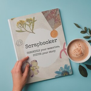 May include: A scrapbook with the word "Scrapbooker" and the text "Chronicle your memories, STITCH your story" is open on a light blue surface. The cover features floral and decorative elements. A cup of coffee and a sprig of leaves are next to the book.