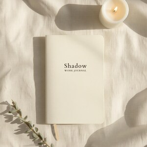 Puede incluir: Un diario de color crema con las palabras "Shadow WORK JOURNAL" impresas en la portada. Una vela blanca encendida y una ramita de vegetación junto al diario sobre una tela color crema.