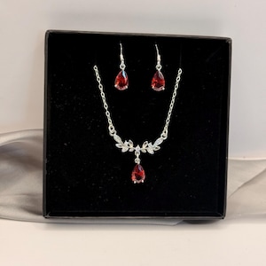 Può includere: Un set di gioielli composto da una collana color argento e orecchini pendenti abbinati. La collana presenta un ciondolo centrale con pietra rossa a goccia, arricchito da dettagli argentati a forma di foglia. Gli orecchini hanno pietre identiche.