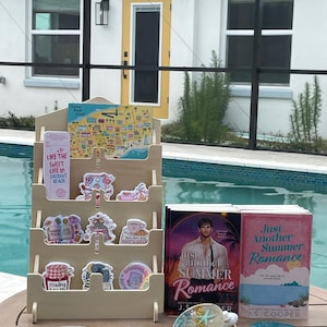 May include: A wooden display stand with books, stickers, and resin coasters. The books are titled "Just Another Summer Romance" by J.S. Cooper. The stickers have various designs, and the coasters feature embedded starfish and glitter. The background includes a swimming pool and a house.