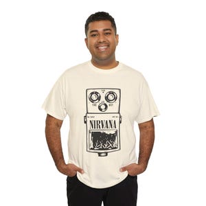 Puede incluir: Una camiseta de manga corta color crema con un estampado gráfico negro de un pedal de efectos para guitarra. El diseño del pedal incluye tres perillas circulares etiquetadas como Time, Level y Dist, con la palabra NIRVANA en letras negras.