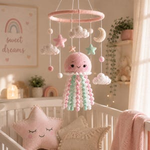 May include: A crochet baby mobile featuring a central pink jellyfish with long, spiraling tentacles in shades of pink, white, and mint green. The mobile includes hanging crochet clouds, stars, and a crescent moon suspended from a circular pink frame.