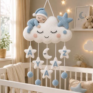 Op de afbeelding: Handgemaakte gehaakte baby mobile met een slapende baby op een witte wolk met blauwe accenten. De mobiel bevat blauwe sterren, een maan en hangende sterren met letters. Het hangt boven een wit babybedje.