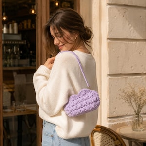 May include: A light purple crochet handbag with a cloud-like shape and a textured popcorn stitch pattern. The bag features a matching thin crochet strap and is worn over the shoulder, paired with a white long-sleeve knit sweater and blue jeans.