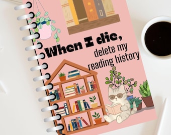 May include: A spiral-bound notebook with a pink cover featuring a hanging plant, a bookshelf, and a cat wearing sunglasses reading a book. The text reads, When I die, delete my reading history. The notebook is positioned on a white surface.