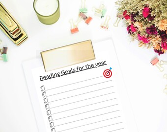 May include: A white clipboard with a gold clip featuring the printed text Reading Goals for the year, a red target icon, and a list of book icons. Surrounding the clipboard are a gold stapler, a green candle, colorful binder clips, and dried flowers.
