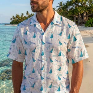 May include: A short-sleeved button-down shirt in white featuring an all-over repeating pattern of the character Elsa from Frozen in a blue dress, interspersed with blue snowflake motifs and the word Frozen printed in blue text throughout.