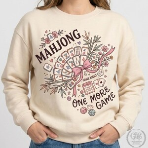 Peut inclure: Un sweat-shirt à col rond couleur crème avec une impression graphique de tuiles de Mahjong disposées en éventail, nouées par un ruban rose. Le motif comprend des tiges de bambou, des fleurs de cerisier, des dés et le texte "MAHJONG" et "ONE MORE GAME".