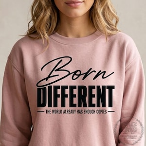 Op de afbeelding: Een oudroze sweater met ronde hals en zwarte tekst die Born DIFFERENT leest en daaronder THE WORLD ALREADY HAS ENOUGH COPIES. Het ontwerp is gecentreerd op de borst en het kledingstuk heeft lange mouwen en een geribbelde halslijn.