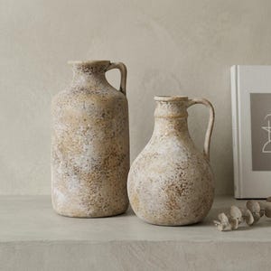 Puede incluir: Dos jarras de cerámica con un acabado texturizado y desgastado en tonos beige, bronceado y crema. La jarra más alta tiene un cuerpo cilíndrico, mientras que la más corta presenta una base redondeada y bulbosa. Ambos recipientes incluyen una asa.