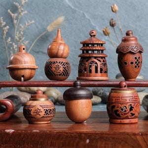 May include: A collection of seven small, carved wooden ornaments featuring various shapes including a bell, a gourd with the character 意, a pagoda, an acorn, a round container with the character 安, a smooth acorn, and a cylindrical jar.