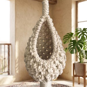 May include: A hanging macrame pod crafted from thick, off-white cotton cord. The structure features an intricate, open-weave knotted pattern with a teardrop shape, suspended from a single thick braided cord at the top, creating a hollow interior.