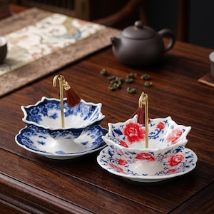 May include: Two ceramic tea strainers with saucers, each featuring a gold-toned metal handle with a small red tassel. One strainer has a blue and white floral pattern, while the other displays a red and blue floral design on a white base.