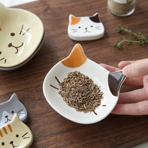 Pode incluir: Um prato de cerâmica branca em forma de gato com detalhes em laranja e castanho escuro nas orelhas, contendo uma pequena quantidade de sementes castanhas. O prato apresenta bigodes pintados e faz parte de um conjunto de loiça felina.