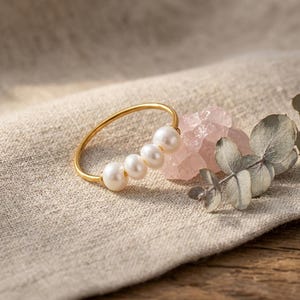 Op de afbeelding: Een dunne goudkleurige ring met vier kleine, ronde, witte parels die op een rij langs de band zijn geplaatst. De ring ligt naast een stuk doorschijnend, lichtroze ruw kristal en een klein takje met gedroogde groene eucalyptusbladeren.