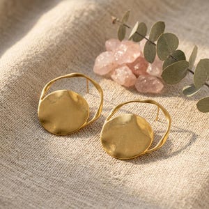 May include: A pair of gold-toned earrings featuring a circular, hammered-texture disc suspended within an open, irregular wire hoop frame. The earrings have a matte finish and are positioned near a cluster of pale pink crystals and green leaves.