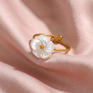 Puede incluir: Anillo dorado con diseño de flor blanca. La flor tiene un centro dorado y pétalos superpuestos. La banda ajustable del anillo tiene dos pequeñas esferas doradas. Joyería de moda.