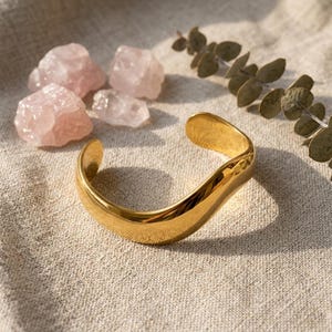 May include: A polished gold-tone cuff bracelet with a smooth, undulating wave-like shape. The open-ended bangle features a reflective metallic surface that catches the light, creating soft highlights and shadows along its curved, continuous form.