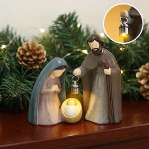 May include: A nativity figurine set featuring three figures in muted tones. One figure wears a blue hooded cloak, another is a bearded figure in a brown robe holding a staff and a small silver lantern, and a third is a small infant in a cradle.
