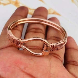 Puede incluir: Un brazalete de metal en tono oro rosa con un mecanismo de cierre central tipo gancho. La banda circular está construida con alambre liso, con detalles decorativos de alambre enrollado que aseguran el área del cierre en ambos lados.