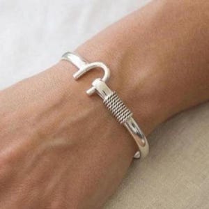 May include: A silver-toned cuff bracelet featuring a hook-and-eye style closure mechanism. The band has a smooth, polished finish on one side and a textured, rope-like twisted metal detail near the clasp, creating a distinct visual contrast.