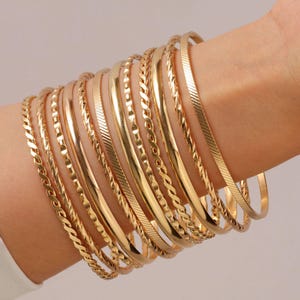 Puede incluir: Una colección de doce brazaletes de metal en tono dorado usados juntos. El conjunto incluye varios diseños como bandas lisas pulidas, texturas de cuerda trenzada y brazaletes con patrones grabados en diagonal, todos con un acabado metálico brillante.