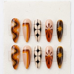May include: A set of ten almond-shaped press-on nails featuring a nude base with various designs in shades of brown, orange, and black. Patterns include wavy lines, geometric blocks, a grid of black diamonds, a sunburst motif, and starbursts.