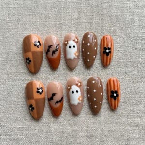May include: A set of ten almond-shaped press-on nails featuring a brown and tan color palette. Designs include a checkered pattern with black flowers, black bats on a tan base, a white ghost with small flowers, brown with white dots, and stripes.