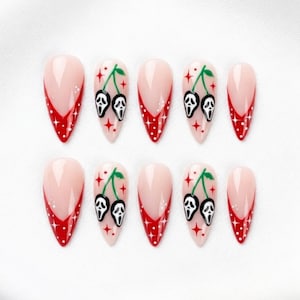 May include: A set of ten almond-shaped press-on nails featuring a nude base with red French tips. Some nails display a cherry-like design where the fruit is replaced by white and black ghost masks with green stems, accented by small red star shapes.