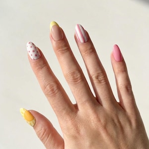K&ouml;nnte beinhalten: Mandelformige Fingern&auml;gel mit verschiedenen Nageldesigns. Der Daumen zeigt ein gelbes Sonnenmotiv, der Zeigefinger hat rosa Punkte, der Mittelfinger eine gelbe French-Spitze, der Ringfinger rosa Streifen und der kleine Finger ist einfarbig rosa.