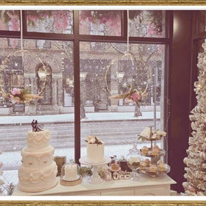 May include: A wedding display with a three-tiered cake, smaller cakes, and desserts. Two decorated Christmas trees are on either side. A snowy street scene is visible through the window, creating a festive atmosphere.