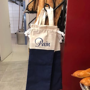 Peut inclure: Un sac fourre-tout en toile crème et bleu marine avec le mot "Pain" en écriture cursive. Le sac est suspendu dans une boulangerie, avec plusieurs pains en arrière-plan. Idéal pour transporter du pain.