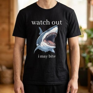 May include: Black t-shirt featuring a shark illustration with an open mouth, displaying sharp teeth. The text "watch out" is above the shark, and "i may bite" is below. The shark is colored in shades of blue and white.