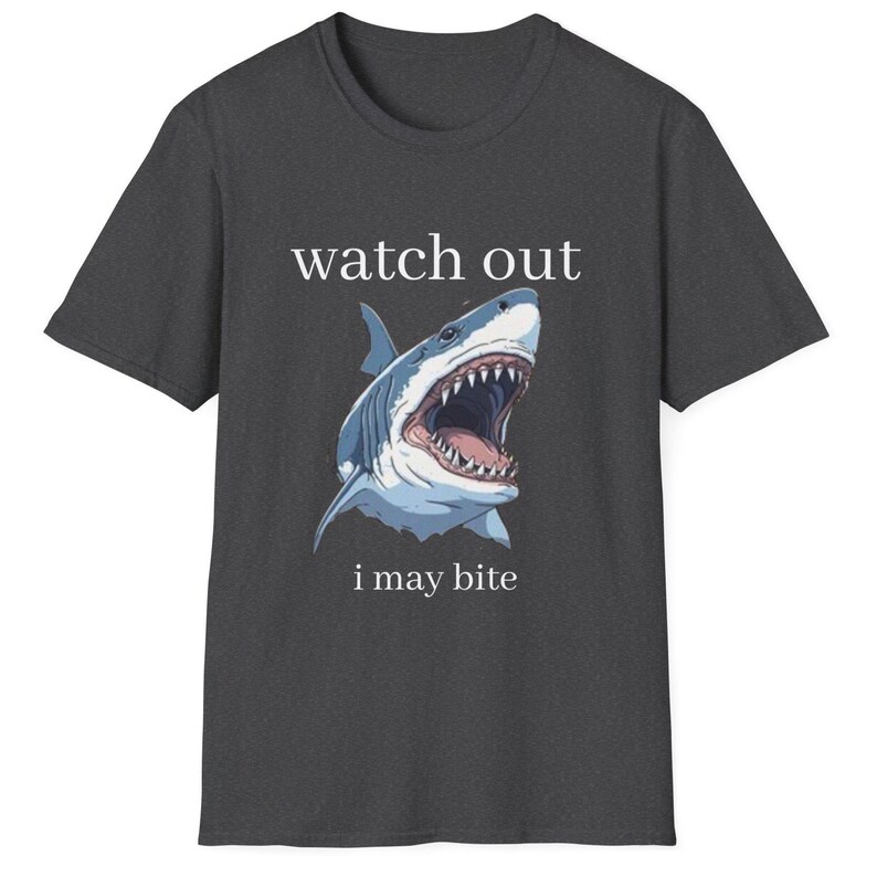 May include: Dark gray t-shirt featuring a graphic of a shark with an open mouth, teeth visible. The text "watch out" and "i may bite" are above and below the shark illustration. A casual tee.