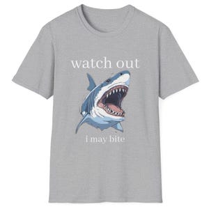 May include: A gray t-shirt featuring a shark illustration with an open mouth, displaying sharp teeth. The text "watch out" is above the shark, and "i may bite" is below. The shark is blue and white.