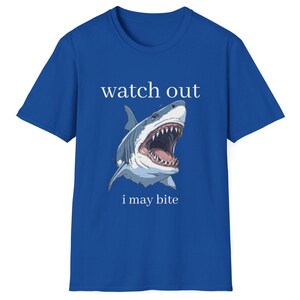 May include: Royal blue t-shirt featuring a detailed illustration of a shark with an open mouth, teeth visible. The text "watch out" is above the shark, and "i may bite" is below.