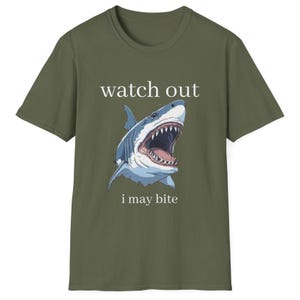 May include: Olive green t-shirt featuring a graphic of a shark with an open mouth, teeth visible. The text "watch out" is above the shark, and "i may bite" is below. The shirt is a crew neck.