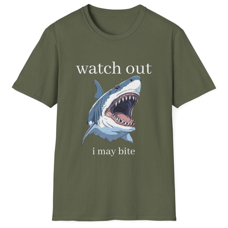 May include: Olive green t-shirt featuring a graphic of a shark with an open mouth, teeth visible. The text "watch out" is above the shark, and "i may bite" is below. The shirt is a crew neck.
