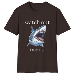May include: A brown t-shirt featuring a detailed illustration of a shark with an open mouth, displaying sharp teeth. The text "watch out i may bite" is printed above and below the shark image.